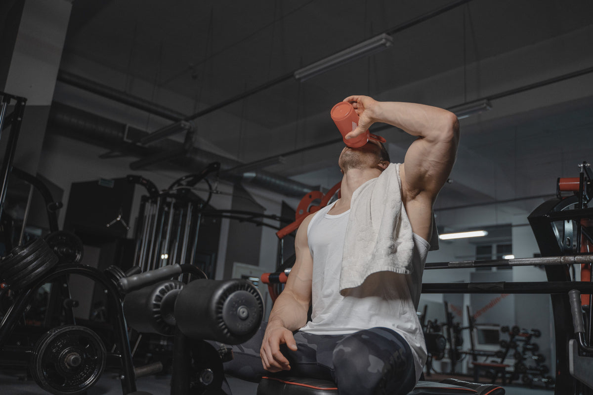 Man in a white tank top drinking a protein shake in a gym after a workout
